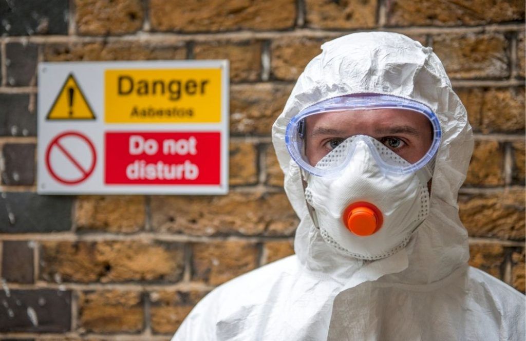 Person is hazmat suit in front of asbestos sign on wall | Featured image for can I remove asbestos myself blog from Asbestos Removals Brisbane.